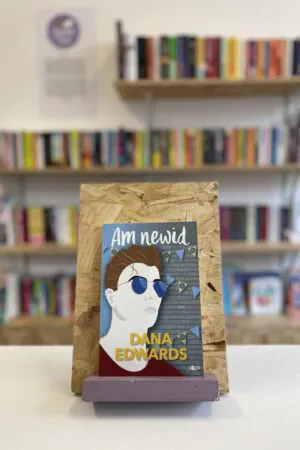 A copy of 'Am Newid' sits on a stand in front of multiple shelves of other books.