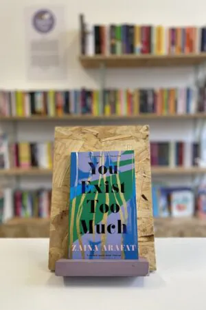 Cymraeg: Copi o 'You Exist Too Much' yn sefyll ar stondin llyfrau, tu blaen silffoedd o lyfrau yn y cefndir. | English: A copy of 'You Exist Too Much' sits on a stand in front of multiple shelves of other books.