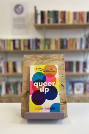 Cymraeg: Copi o 'Queer Up' yn sefyll ar stondin llyfrau, tu blaen silffoedd o lyfrau yn y cefndir. | English: A copy of 'Queer Up' sits on a stand in front of multiple shelves of other books.