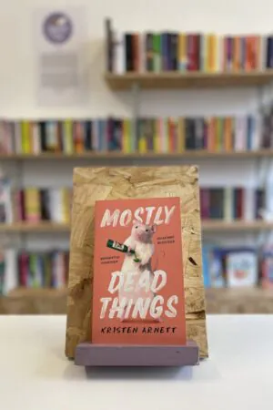 A copy of 'Mostly Dead Things' sits on a stand in front of multiple shelves of other books.