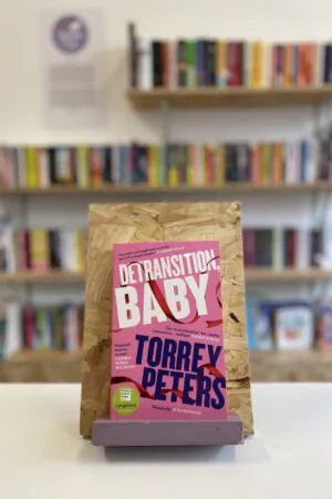 Cymraeg: Copi o 'Detransition, Baby' yn sefyll ar stondin llyfrau, tu blaen silffoedd o lyfrau yn y cefndir. | English: A copy of 'Detransition, Baby' sits on a stand in front of multiple shelves of other books.