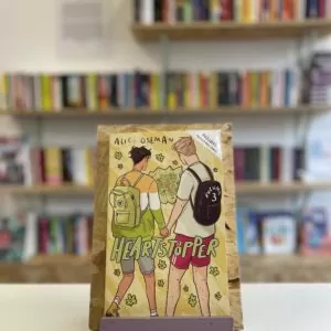 Cymraeg: Copi o 'Heartstopper vol. 3' yn sefyll ar stondin llyfrau, tu blaen silffoedd o lyfrau yn y cefndir. | English: A copy of 'Heartstopper vol. 3' sits on a stand in front of multiple shelves of other books.