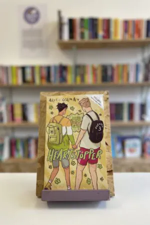 Cymraeg: Copi o 'Heartstopper vol. 3' yn sefyll ar stondin llyfrau, tu blaen silffoedd o lyfrau yn y cefndir. | English: A copy of 'Heartstopper vol. 3' sits on a stand in front of multiple shelves of other books.