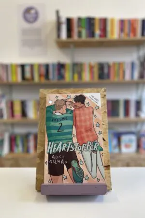 Cymraeg: Copi o 'Heartstopper vol. 2' yn sefyll ar stondin llyfrau, tu blaen silffoedd o lyfrau yn y cefndir. | English: A copy of 'Heartstopper vol. 2' sits on a stand in front of multiple shelves of other books.