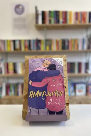 Cymraeg: Copi o 'Heartstopper vol. 4' yn sefyll ar stondin llyfrau, tu blaen silffoedd o lyfrau yn y cefndir. | English: A copy of 'Heartstopper vol. 4' sits on a stand in front of multiple shelves of other books.
