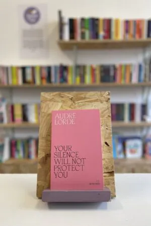 A copy of 'Your Silence Will Not Protect You' sits on a stand in front of multiple shelves of other books.