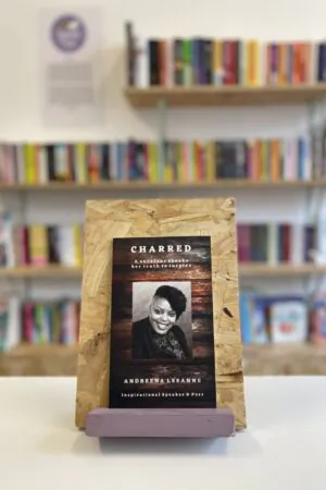 Cymraeg: Copi o 'Charred' yn sefyll ar stondin llyfrau, tu blaen silffoedd o lyfrau yn y cefndir. | English: A copy of 'Charred' sits on a stand in front of multiple shelves of other books.