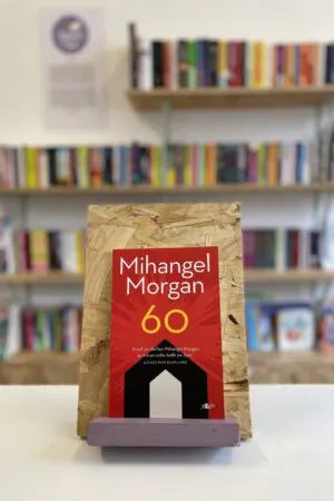 Cymraeg: Copi o '60' yn sefyll ar stondin llyfrau, tu blaen silffoedd o lyfrau yn y cefndir. | English: A copy of '60' sits on a stand in front of multiple shelves of other books.