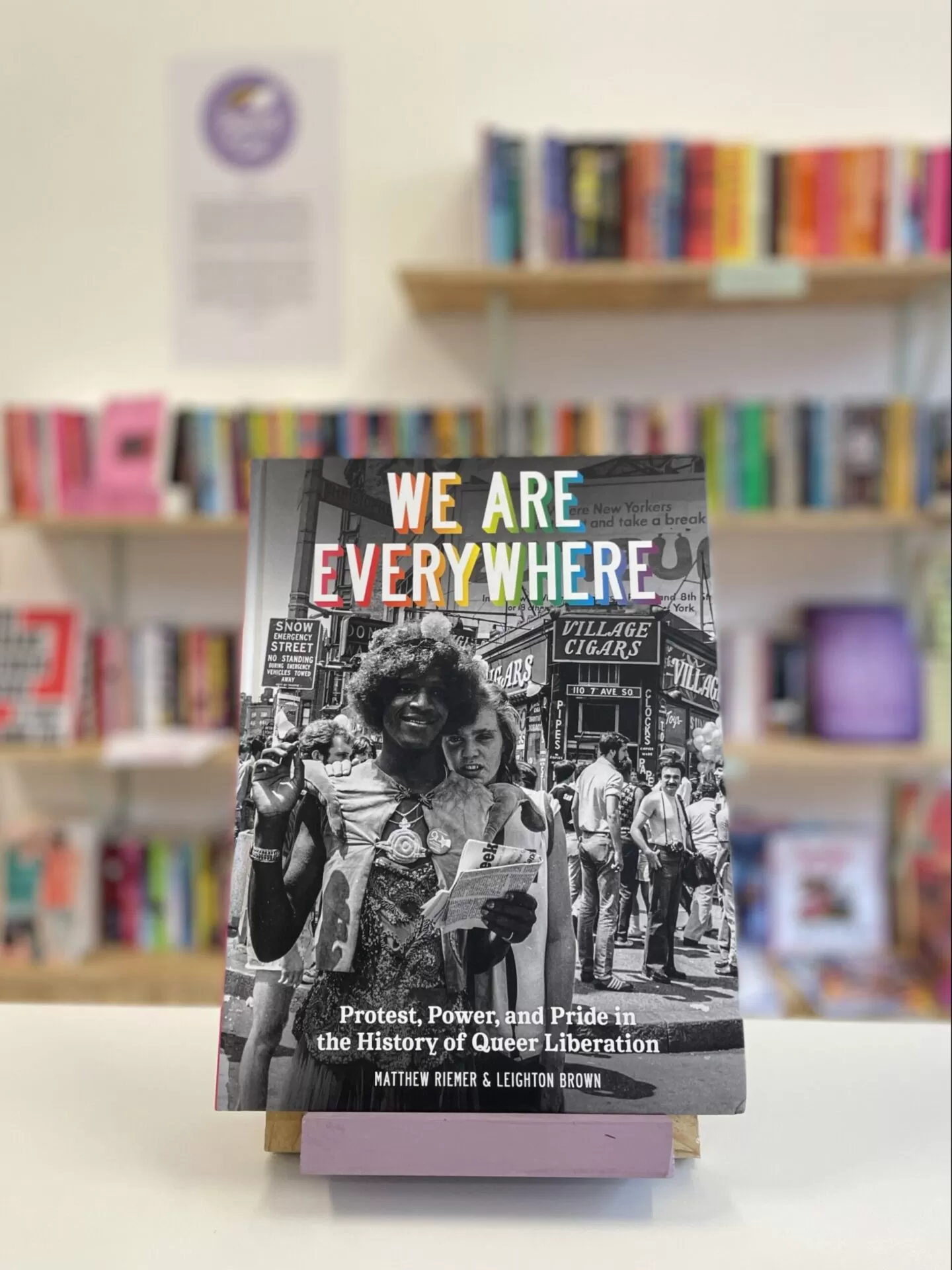 Cymraeg: Copi o 'We Are Everywhere' yn sefyll ar stondin llyfrau, tu blaen silffoedd o lyfrau yn y cefndir. | English: A copy of 'We Are Everywhere' sits on a stand in front of multiple shelves of other books.