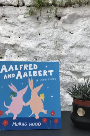 On a black table, a copy of 'Aalfred and Aalbert' by Morag Hood stands on a black table, next to a potted plant.  Behind them, a stone, white wall obscures the background.