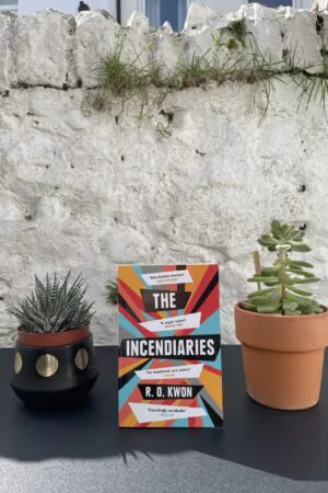 On a black table, between two potted plants, a copy of the book is standing, with a white, stone wall in the background.