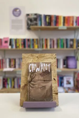 Cymraeg: Copi o 'Chwant' yn sefyll ar stondin llyfrau, tu blaen silffoedd o lyfrau yn y cefndir. | English: A copy of 'Chwant' sits on a stand in front of multiple shelves of other books.
