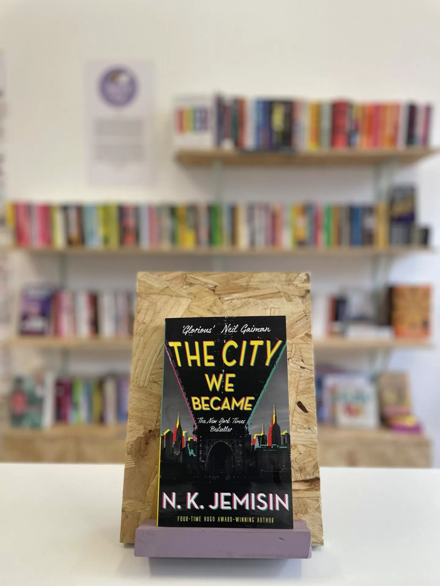 Cymraeg: Copi o 'The City We Became' yn sefyll ar stondin llyfrau, tu blaen silffoedd o lyfrau yn y cefndir. | English: A copy of 'The City We Became' sits on a stand in front of multiple shelves of other books.