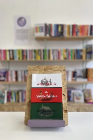 Cymraeg: Copi o 'Mês Bach a Gwreiddiau' yn sefyll ar stondin llyfrau, tu blaen silffoedd o lyfrau yn y cefndir. | English: A copy of 'Mês Bach a Gwreiddiau' sits on a stand in front of multiple shelves of other books.