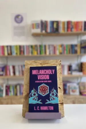 Cymraeg: Copi o 'Melancholy Vision' yn sefyll ar stondin llyfrau, tu blaen silffoedd o lyfrau yn y cefndir. | English: A copy of 'Melancholy Vision' sits on a stand in front of multiple shelves of other books.