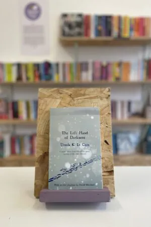 Cymraeg: Copi o 'The Left Hand of Darkness' yn sefyll ar stondin llyfrau, tu blaen silffoedd o lyfrau yn y cefndir. | English: A copy of 'The Left Hand of Darkness' sits on a stand in front of multiple shelves of other books.