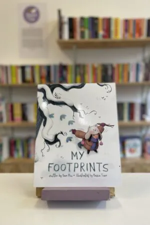 Cymraeg: Copi o 'My Footprints' yn sefyll ar stondin llyfrau, tu blaen silffoedd o lyfrau yn y cefndir. | English: A copy of 'My Footprints' sits on a stand in front of multiple shelves of other books.