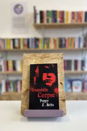 Cymraeg: Copi o 'Exquisite Corpse' yn sefyll ar stondin llyfrau, tu blaen silffoedd o lyfrau yn y cefndir. | English: A copy of 'Exquisite Corpse' sits on a stand in front of multiple shelves of other books.