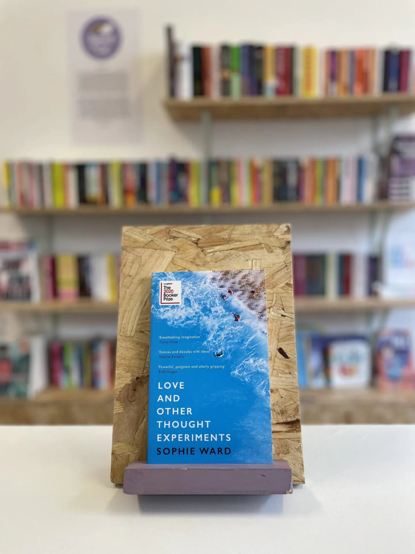 Cymraeg: Copi o 'Love and Other Thought Experiments' yn sefyll ar stondin llyfrau, tu blaen silffoedd o lyfrau yn y cefndir. | English: A copy of 'Love and Other Thought Experiments' sits on a stand in front of multiple shelves of other books.