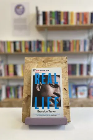 Cymraeg: Copi o 'Real Life' yn sefyll ar stondin llyfrau, tu blaen silffoedd o lyfrau yn y cefndir. | English: A copy of 'Real Life' sits on a stand in front of multiple shelves of other books.
