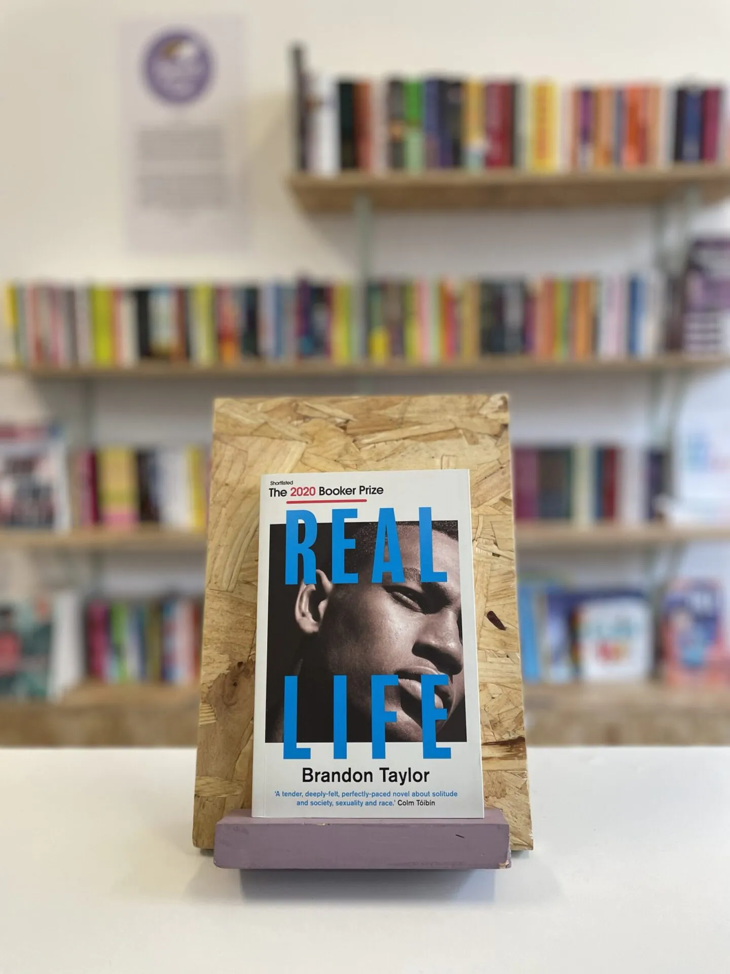 Cymraeg: Copi o 'Real Life' yn sefyll ar stondin llyfrau, tu blaen silffoedd o lyfrau yn y cefndir. | English: A copy of 'Real Life' sits on a stand in front of multiple shelves of other books.