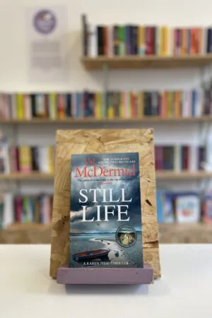 Cymraeg: Copi o 'Still Life' yn sefyll ar stondin llyfrau, tu blaen silffoedd o lyfrau yn y cefndir. | English: A copy of 'Still Life' sits on a stand in front of multiple shelves of other books.