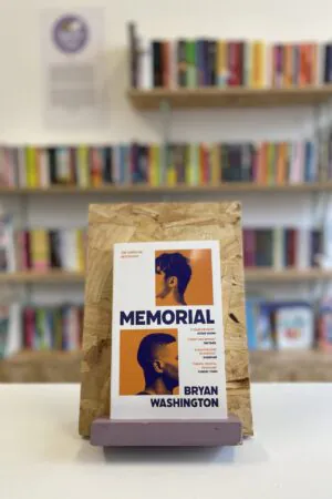 Cymraeg: Copi o 'Memorial' yn sefyll ar stondin llyfrau, tu blaen silffoedd o lyfrau yn y cefndir. | English: A copy of 'Memorial' sits on a stand in front of multiple shelves of other books.