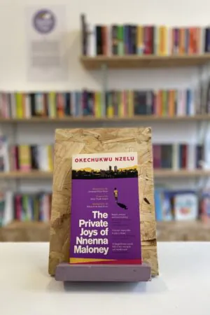 A copy of 'The Private Joys of Nnenna Maloney' sits on a stand in front of multiple shelves of other books.