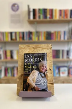 Cymraeg: Copi o 'In My Mind's Eye' yn sefyll ar stondin llyfrau, tu blaen silffoedd o lyfrau yn y cefndir. | English: A copy of 'In My Mind's Eye' sits on a stand in front of multiple shelves of other books.