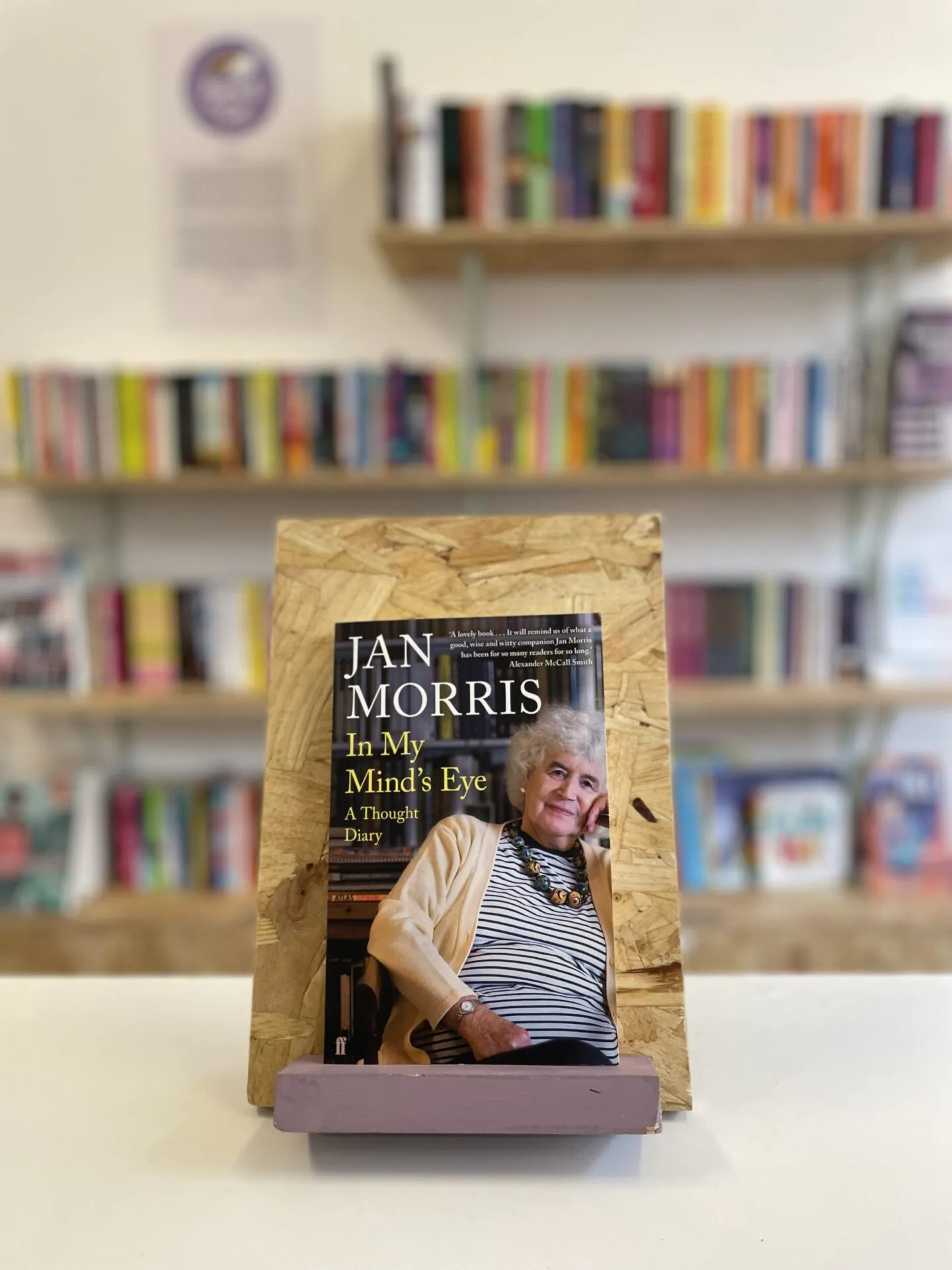 Cymraeg: Copi o 'In My Mind's Eye' yn sefyll ar stondin llyfrau, tu blaen silffoedd o lyfrau yn y cefndir. | English: A copy of 'In My Mind's Eye' sits on a stand in front of multiple shelves of other books.