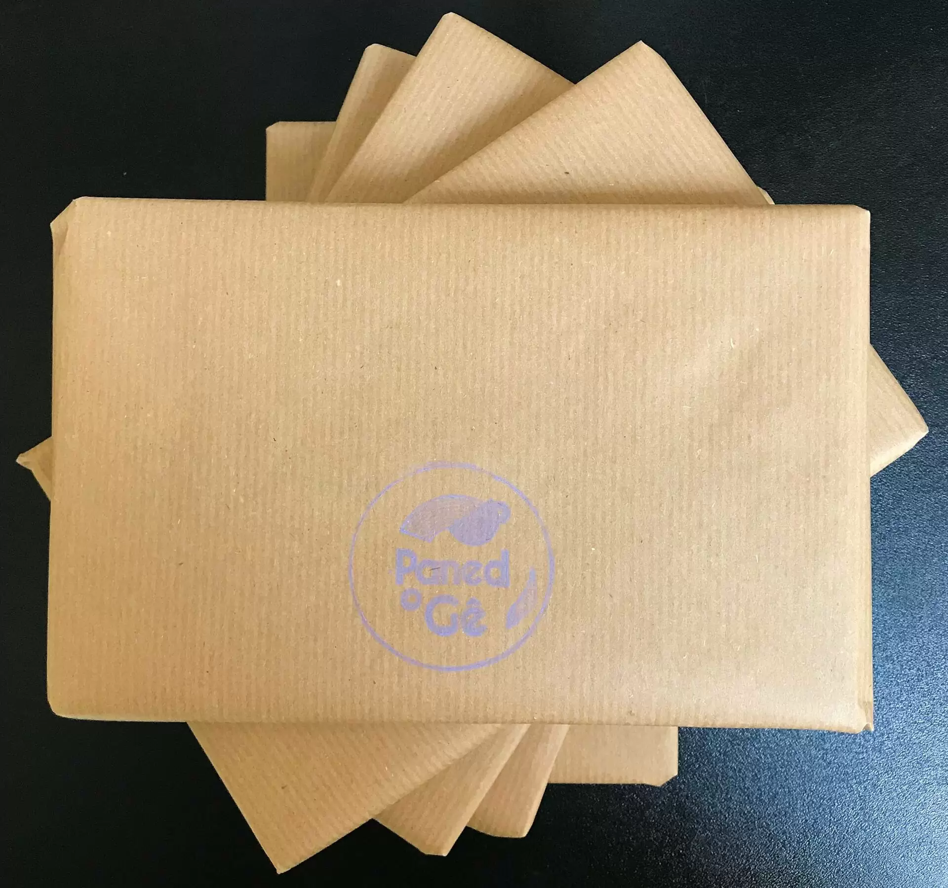 A pile of brown, square parcels on a black surface, asymmetrically arranged. Paned o Gê's logo is printed in lavender on the top parcel.